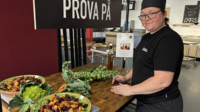 Svartklädd leende kvinnlig kock vid bord med färgglada grönsaker.