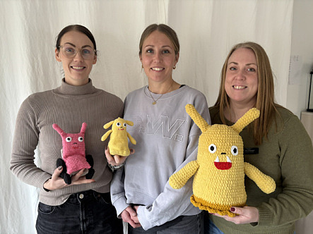 Tre glada kvinnor håller upp två gula och en rosa virkad firgur.