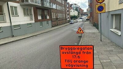 Röd skylt med text att gatan stängs av från 17/6. Tom gata och tegelhus bakom skylten.