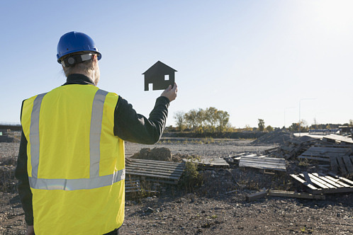 Byggarbetare iklägg gul reflexväst står på en byggarbetsplats med ryggen mot kameran och håller upp en hus-siluett skylt i trä.