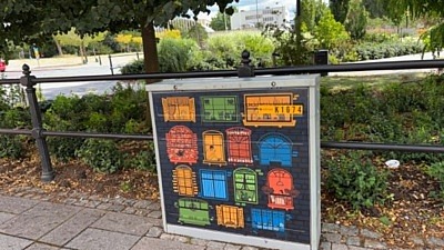 Ett grått elskåp vid en park med ett konstverk som föreställer ritade fönster i olika färger
