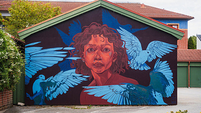 Färgstark muralmålning med ett stort porträtt målat över en hög husfasad. Konstverket använder starka färger och mjuka övergångar som skapar ett uttrycksfullt och levande motiv.