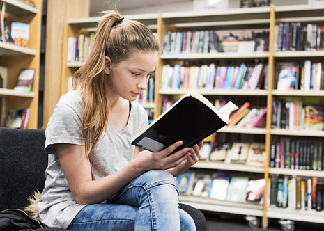 En ung tjej sitter i ett bibliotek och läser en bok