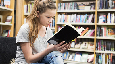 En ung tjej sitter i ett bibliotek och läser en bok